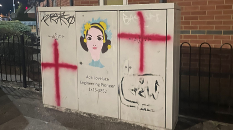 A grey utility box, with St George's crosses on the right side and left side, and the cartoon of Ada Lovelace, which features a woman with brown hair, a yellow headdress and underneath: "Ada Lovelace, engineering pioneer, 1815-1852". 