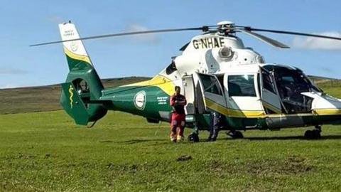 A green, yellow and white helicopter is sat on a large patch of grass. Great North Air Ambulance Service members wait outside the helicopter. 