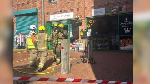 An emergency response scene outside a row of brick shopfronts on a pedestrian street. Four firefighters are in the centre of the scene, wearing full protective gear (tan fire-resistant suits, helmets, and high‑visibility markings). Fire hoses are laid out on the ground, looping across the brick‑paved walkway and leading toward the buildings. Red and white safety tape cordons off the area in the foreground.