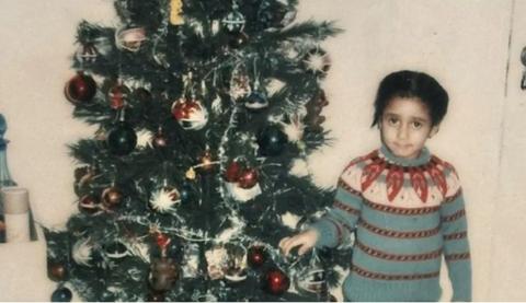 A child stands next to a Christmas tree. She has dark hair and dark eyes, and is wearing a red and blue striped jumper