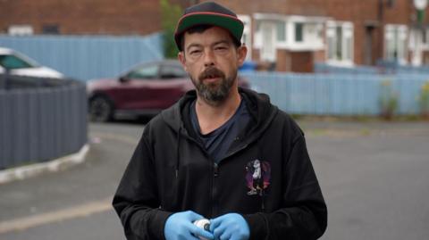 Martin Withers, who is wearing a black hooded jacket and a baseball cap, is staring directly at the camera. He is holding a can of spray paint and is wearing protective blue gloves.