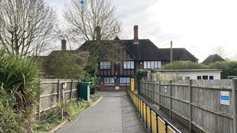 An external view of the school taken from a driveway leading up to the mock-Tudor building. A wooden fence can be seen on either side of the image, with plants on the left and a yellow barrier on the right.