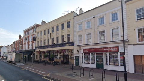 Image taken from the street looking towards the Golden Cross Hotel which is three storeys tall with large square windows. The façade is a pale yellow and above the entrance is a large blue sign with gold writing that reads Golden Cross Hotel. 