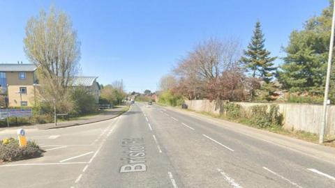 A Google Streetview of Bristol Road, which runs through part of Chippenham. There are a number of trees and bushes running alongside the road, with a fence to the right and what appears to be an office to the left. A housing estate is visible in the distance. 