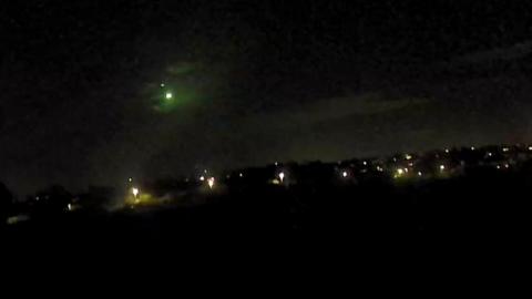 A light can be seen in the black night sky. There are street lights on the ground.