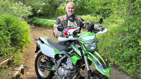 A man sits on a dirt bike on a dirt path. He is bald and he has a grey beard. He is wearing a black and brown jacket. The bike is lime green. 