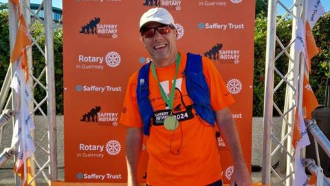 Hayden Davies at the finish line of the 2024 event. He is grinning and wearing a gold-coloured medal with a green ribbon around his neck. He is wearing an orange T-shirt which is printed with: Saffery Rotary Walk 2024, sunglasses and a cap. Behind him is a banner with Saffery Trust, Rotary in Guernsey and Saffery Rotary Walk logos and branding on it.