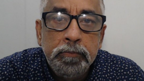 Vijay Chawla, wearing a dark blue shirt with white speckles, and with a grey beard and glasses, looks straight ahead in his police mugshot