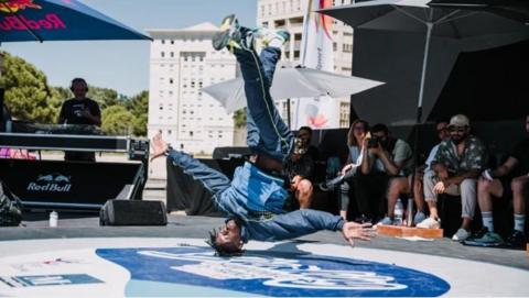 A young man wearing a blue tracksuit spins on his head while a DJ stands under an umbrella in the background and people gather round to watch. It is a sunny day