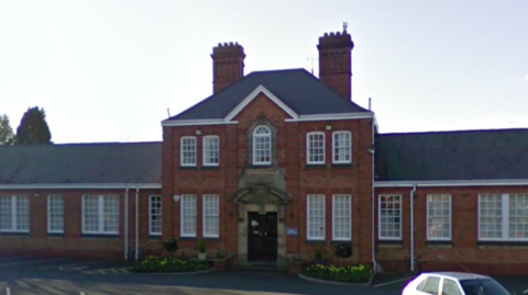 The front of a red brick school with several white framed windows on either side of an entrance way with a large black door