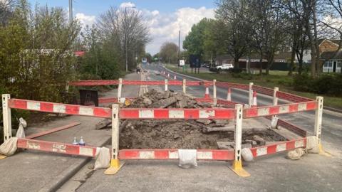 A large hole in the ground around the burst water main with orange and white safety barriers around it