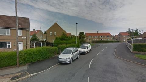An asphalt road with white line markings leads to a series of council house. Two cars, one white, the other silver are parked straddling the road and pavement.