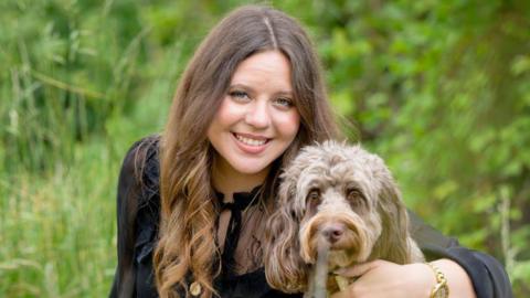 Devon Taverner-Hailou has long brown hair and is wearing a black top while smiling. Her left arm is around Molly, a light brown cockapoo, sitting next to her. 