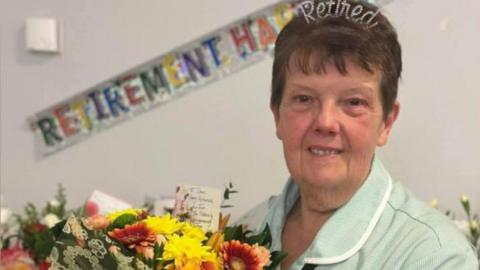 A woman holds a bunch of yellow and orange flowers. She has short brown hair and she is wearing a tiara that says "retired" on it. She is wearing a pale green uniform. A banner saying "RETIREMENT" in capital letters can be seen in the background.