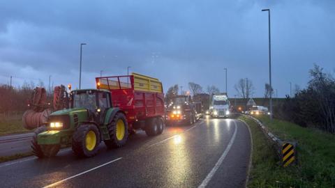 A tractor and trailer lead a slow-moving protest on a roundabout in County Tyrone on a wet evening. The green tractor is pulling a red trailer. It is followed by a second tractor. There is a large white HGV in the outer lane and a queue of traffic behind