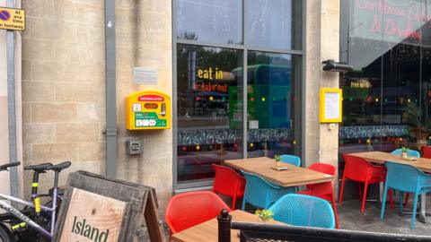 A yellow defibrillator is pictured on the exterior wall of a bar. There are tables and chairs, in red and blue, in front of two large plate-glass windows.