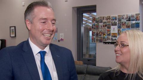 Paul Givan in blue suit, white shirt and blue tie, smiling and laughing alongsideand blonde haired woman wearing glasses in youth centre, photos on wall behind and a sofa