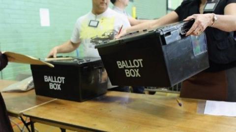 People placing two black ballot boxes on a trestle table at an election count
