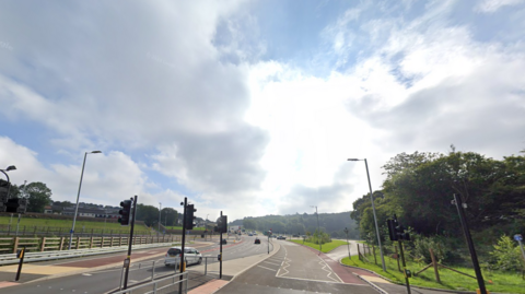 Forder Valley Road in Plymouth. The photo is taken by a set of traffic lights before the next set of traffic lights. There are three lanes with a white car in the middle.