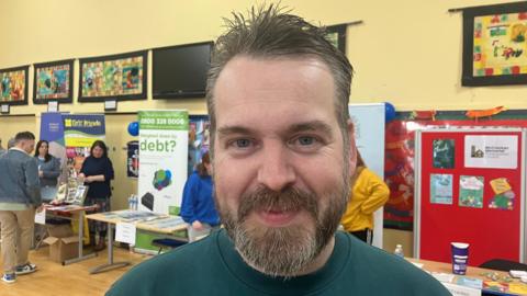 A bearded man wearing a green sweater looks directly at the camera, he has a smile on his face