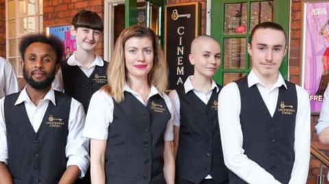Five people are standing outside a building with two green doors, one of which is open. They are wearing black-and-white Lockworks Cinema uniforms that include its name.