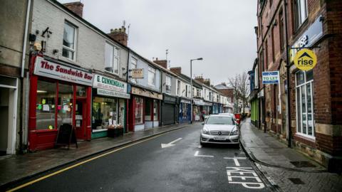 Crook town centre. A sandwich shop is one of many shops lining a high street made up of a row of terraced buildings. Cars are parked on the other side of the road.