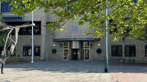 Entrance to Bradford Crown Court - a stone building with a revolving door. In the front of the picture is a tree and also part of a sculpture which sits outside the front of the building.