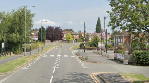 A suburban street lined by estate housing with trees either side and a pedestrian crossing visible