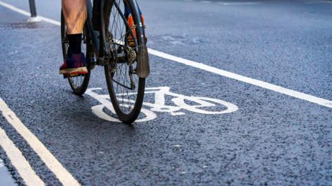 A cyclist riding in a designated bike lane marked by white lines and a painted bicycle symbol on a road. Only the bike’s rear wheel and the rider’s legs and shoes are visible.