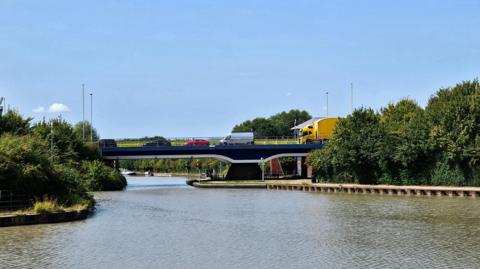 A bridge carries traffic, including a bright yellow lorry, a van, and three cars, over a canal on a sunny summer's day. There are trees on either side of the canal and canal boats can be seen in the distance on the other side of the bridge. There are hardly any clouds in the sky.