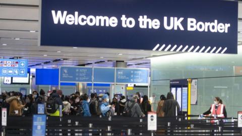 People queue at an airport with a sign reading "welcome to the UK border" hanging above them.