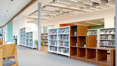 An empty modern looking library with rows and rows of books arranged in white and brown bookcases. A chair and tables are to the right 