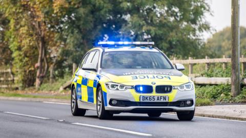 A police car with blue lights on is seen travelling on a road. It has lettering on it which reads 'ANPR interceptor' and reverse lettering which says 'police'.