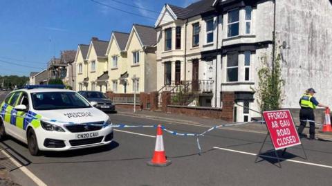 A police car is seen along a road, with police tape and a red "Road Closed" sign
