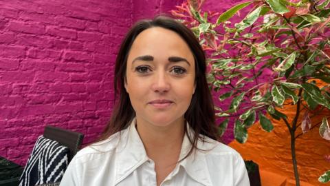 Danielle pictured looking into the camera with a neutral expression; she's wearing a white collared shirt. She's sat in one of the restaurant's outside areas, which has its walls painted bright pink and orange. Behind her, to her left is a small tree with green leaves. To her right is a black table and chairs with a black and white chevron-patterned cushion. 
