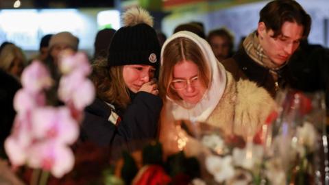 Two young women hug each other while one cries, laying floral tributes