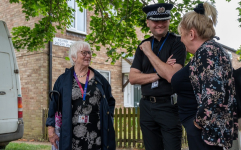 A woman in a black floral dress speaks to a police officer and another woman in a blue floral dress in the middle of a housing estate. 