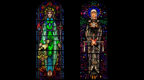 Two stained glass windows, each featuring a Catholic saint. The one on the left has their hand on a lamb. The one on the right is holding a crucifix.
