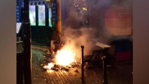 A pile of rubbish in flames outside of a shop on Corn Street in Bristol. The picture has been taken at night.