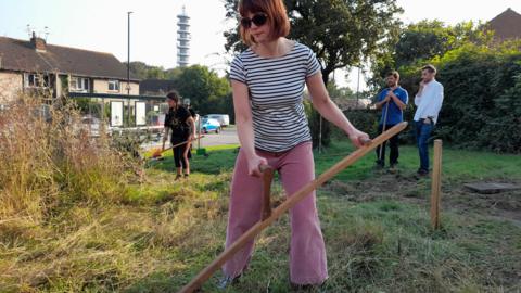 Four people are working together to scythe grass in Green Meadow. In the foreground, a woman dressed in pink trousers and a black-and-white striped T-shirt is using a garden tool. Behind her, two men are standing and another woman is also at work scattered across the partially-cleared meadow. Piles of cut grass are visible on one side, while a  rows of houses frames the background.