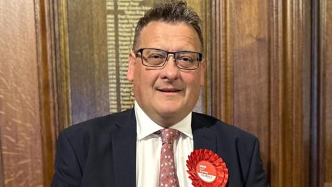 Karl Turner the succesful Labour candidate for Hull East pictured on election night in 2024 wearing a dark suit jacket, white shirt, and patterned pink tie, standing indoors against a wooden paneled wall. A large red rosette with the word ‘Labour’ and smaller text is pinned to the left side of the jacket. 