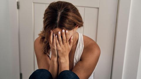 Stock image of an unidentifiable woman sitting on the floor by the door, crying and covering her face with her hands.