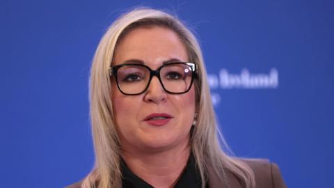 Michelle O'Neill. She has shoulder-length blonde hair, wearing glasses and a brown blazer. The is a blue background.