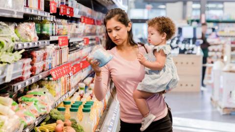 A woman checks the price of an item in a grocery store while holding a baby