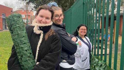 Three young women stand holding a roll of faux shrubbery next to a green metal fence. They are wearing coats and smiling.