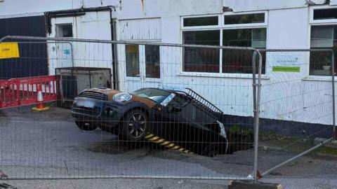 A Mini car's rear has fallen into a hole about 2m deep. Metal fencing has been put up around the area, which is a car park behind a bakery in Redruth.