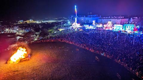 The photograph shows a large crowd watching the 2023 bonfire. The crowd are held back by barriers and behind them you can see fairground rides.