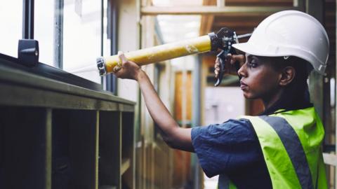 A woman is inside a building and is wearing a white helmet, blue T-shirt and yellow and silver fluorescent jacket. She is holding a caulk gun which has a yellow body and a long white nozzle.