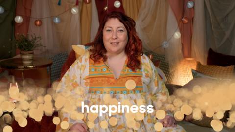 A female presenter in a homely room with lights and the word 'happiness' as a caption.