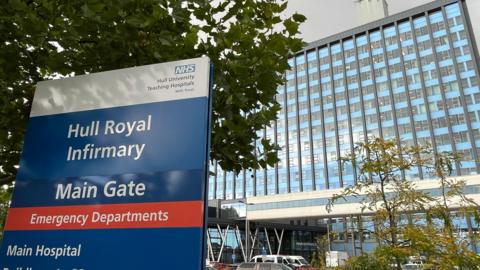 In the foreground, there is a sign that says Hull Royal Infirmary Main Gate and emergency departments. The large oblong hospital building with its blue grey uniformed glass windows stands behind the sign.